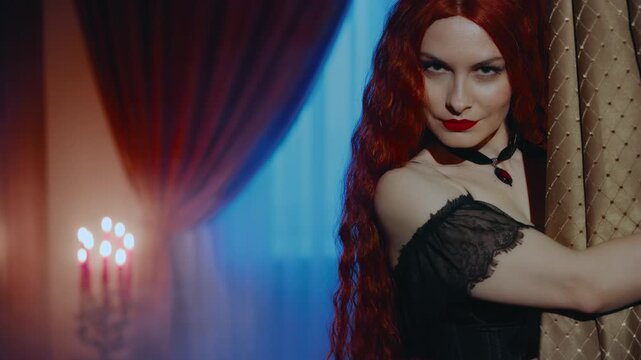 Redhead medieval witch beauty gothic woman hand opens curtain, evil eyes looking into camera. Dark night room. Fantasy lady girl sexy face evening makeup lips long red hai black dress old style art
