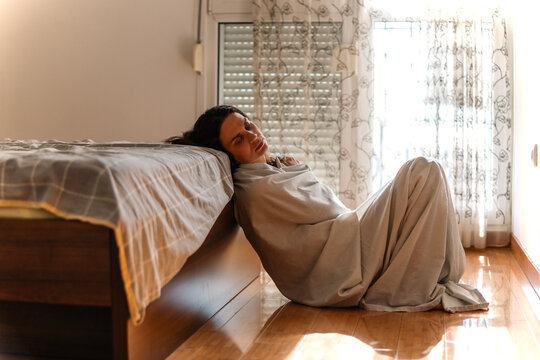 Sad lonely female feeling down, sitting on bedroom floor wrapped in bedsheet. Apathy, grief and despair.