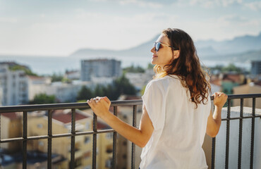 Fototapeta premium Beautiful young woman standing on apartment balcony and enjoying sunny summer day. City view background.