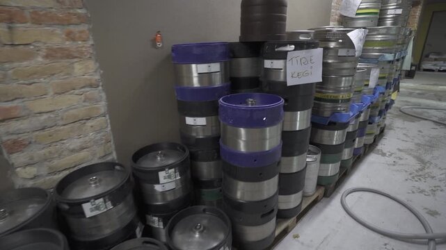 Camera moves past stacked metal beverage kegs with handwritten labels and blue carriers inside an industrial storage room, revealing rows of containers