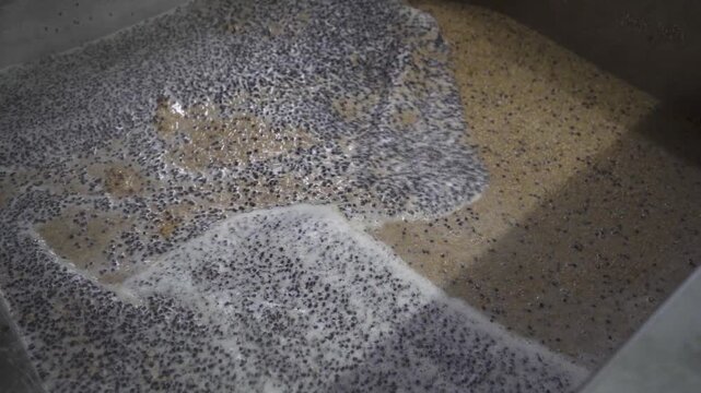 Close up of malt grains being stirred in hot water with a metal paddle, foam moving across the surface during the brewing process in an industrial vessel