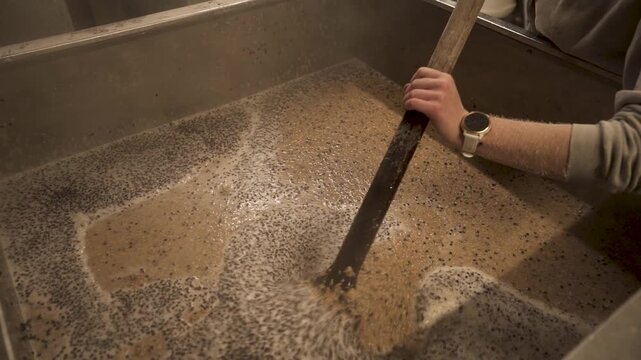 Person stirring a foamy malt mixture in hot water inside a stainless steel brewing vessel during beer production