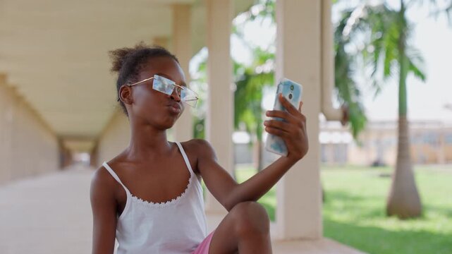 Young girl sits outdoors on concrete steps holding and using her blue cased smartphone to take photos. Girl wears white tank top with lace trim and pink shorts under covered walkway of what