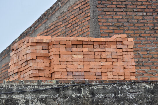 A large stack of red clay bricks sitting on a concrete wall structure at a building construction site against a brick wall.