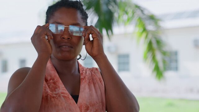 Woman in light outdoor setting holds and puts on rimless eyeglasses while wearing sleeveless pink patterned wrap dress with large hoop earrings. background shows blurred greenery