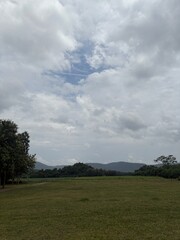 Naklejka premium Open Grass Field Under Cloudy Sky with Trees and Distant Hills