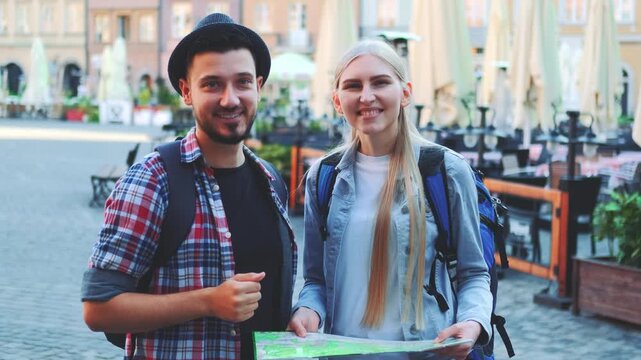 Two young tourists holding a map and smiling while standing in a lively European city square with outdoor cafes and traditional buildings.