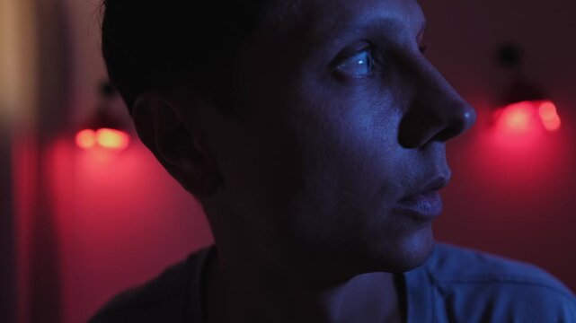 Anxious man looking down and suddenly turning toward a possible threat, experiencing anticipatory fear and dread despite no visible danger in a dark red and blue low key bedroom