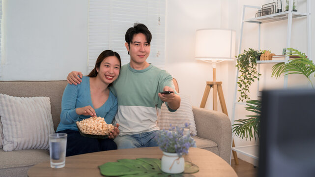 Young Asian couple watching TV eating popcorn together on sofa in cozy living room