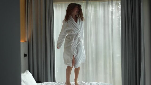 Joyful young woman in a white bathrobe jumping on a bed and having fun in a cozy hotel room.
