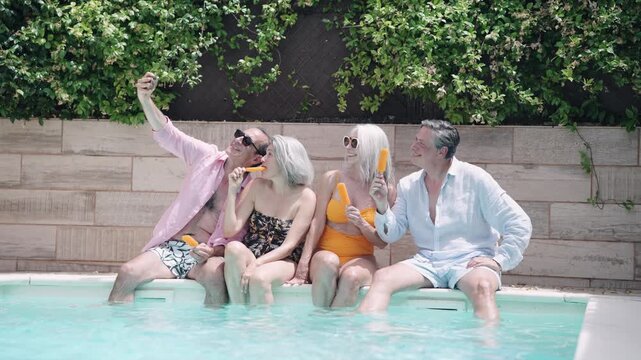 Group of smiling mature friends enjoying their vacation. Cheerful people sitting on the poolside eating popsicles and taking a selfie