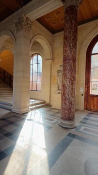 Vertical view of a red marble column with a carved capital and a bright sunbeam on the patterned stone floor of a quiet neoclassical entrance hall, Paris, France