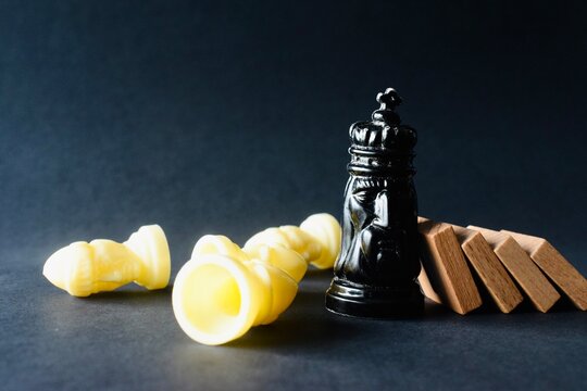 Black king chess piece standing beside falling wooden domino blocks and defeated white pawns on a dark background.