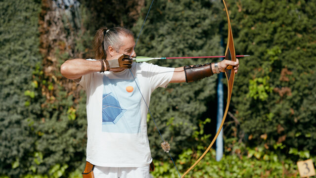 Mature man focusing and aiming a bow and arrow during an outdoor archery session, symbolizing precision and goal achievement