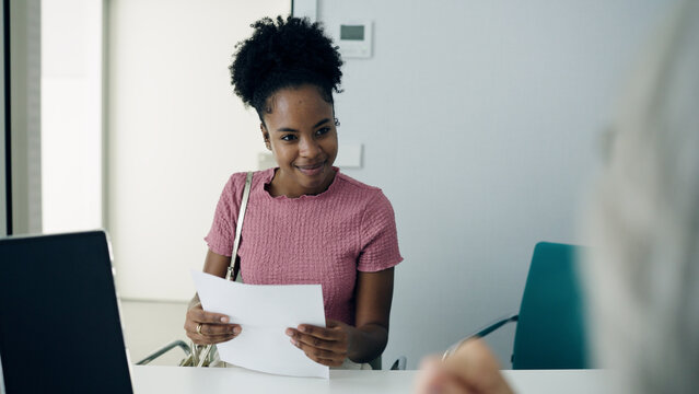 Young African American woman smiling, presenting medical documents to a person at a hospital reception or clinic desk