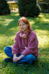 A cute redhead girl sits on grass, looking at the camera while wearing a purple fleece hoodie and pink headphones in a park. © Dmytro
