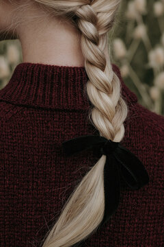 Close rear portrait of a young Caucasian woman with blonde braid, black ribbon, and burgundy knit sweater outdoors.