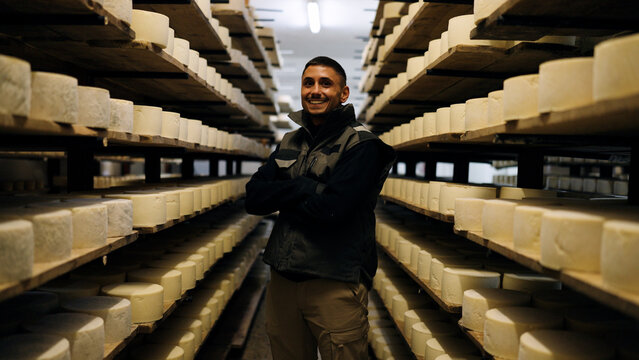 Happy cheesemaker standing with crossed arms in a traditional cheese cellar, surrounded by rows of maturing cheese wheels