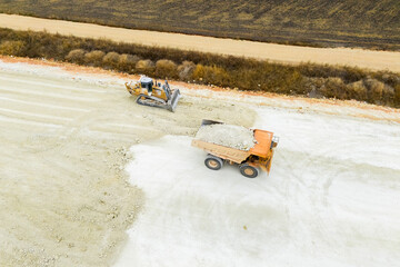 An aerial view of heavy machinery including a dump truck unloading soil, a bulldozer, and a grader working on a road construction embankment. © scharfsinn86