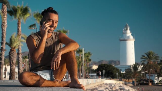 Smiling young man holding a phone to his ear while sitting cross-legged on a concrete seaside ledge near a white lighthouse and palm trees, digital nomad lifestyle, coastal living
