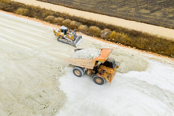 An aerial view of heavy machinery including a dump truck unloading soil, a bulldozer, and a grader working on a road construction embankment. © scharfsinn86