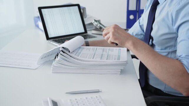 Auditor's hands using a magnifying glass on a stack of financial documents and reports while working at an office desk with a laptop and calculator, focusing on business data verification