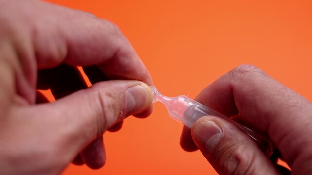 Human hands twisting open a plastic single-use ampoule with saline solution or liquid medicine against a orange background