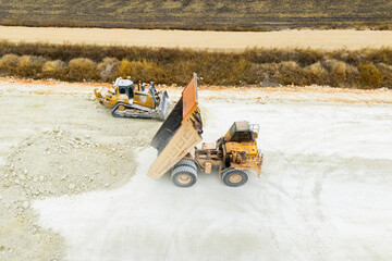 An aerial view of heavy machinery including a dump truck unloading soil, a bulldozer, and a grader working on a road construction embankment. © scharfsinn86