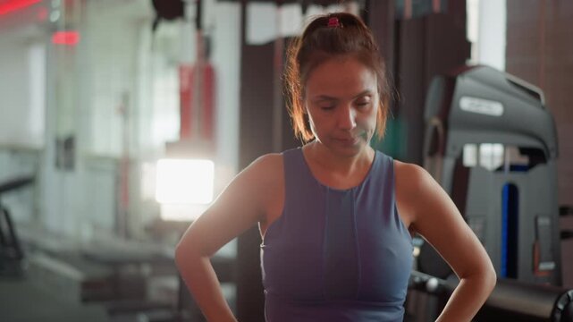 Woman stands in well lit indoor gym hands on hips. Gym equipment like treadmills and weights are visible behind her. Woman looks directly at camera workout