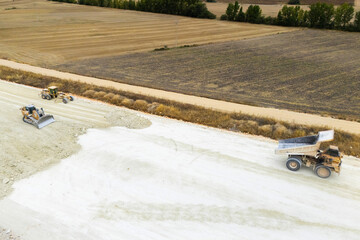 An aerial view of heavy machinery including a dump truck unloading soil, a bulldozer, and a grader working on a road construction embankment. © scharfsinn86