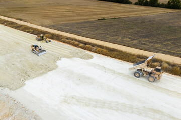 An aerial view of heavy machinery including a dump truck unloading soil, a bulldozer, and a grader working on a road construction embankment. © scharfsinn86