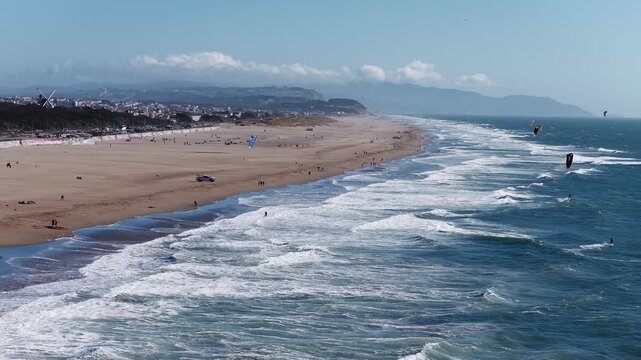 Aerial view tracks along Ocean Beach in San Francisco, California, showing crashing Pacific Ocean surf, sandy shore, kitesurfers riding ocean waves, and the distant Murphy Windmill landmark.