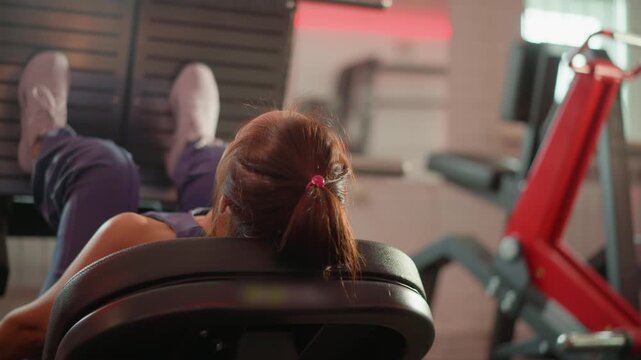 Young girl with brown hair tied back rests her head and legs elevated at end of lower body weight training session. Woman is positioned under red exercise machines wearing casual workout