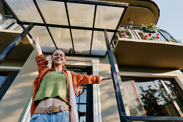 Fototapeta premium Happy young woman with raised arms standing under a glass canopy near her home entrance. Concept of new home, joy, and freedom. Warm sunlight creates a cheerful mood.