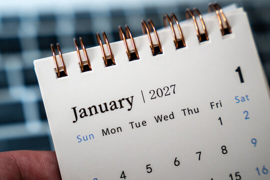 January 2027 calendar held above a blurred laptop keyboard in an office setting, first month of the year with copper spiral binding, concept of business planning, work schedule and annual goals