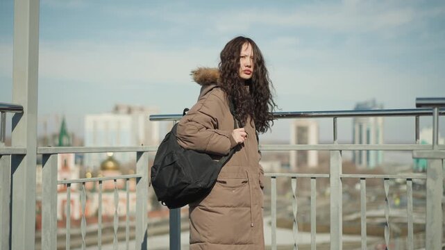 Woman with long curly hair stands on elevated platform wearing brown winter coat over her shoulders. Woman carries black backpack while looking out towards cityscape in daylight. background