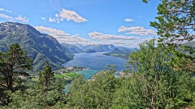 Panoramic view from Nuken Viewpoint overlooking Andalsnes and Romsdalsfjorden. Mountains rise steeply around turquoise waters under bright blue sky. Lush green trees frame foreground Romsdalseggen Rid