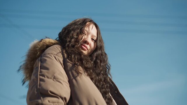 Woman walks outdoors in bright daylight under cloudless blue sky wearing brown winter coat. Her long curly hair moves naturally as woman progresses through scene with power lines and clear