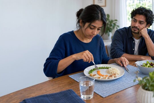 Woman obsessively arranging food on plate with concerned man watching. Eating disorder, food restriction, and mental health concept