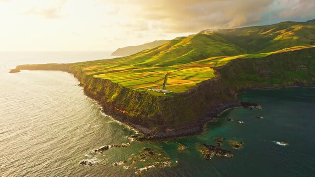Sunrise over Farol do Albarnaz lighthouse on Flores Island, Azores, Portugal. Volcanic coast, patchwork green fields, and Atlantic ocean. Golden hour. Aerial view. Moving forward