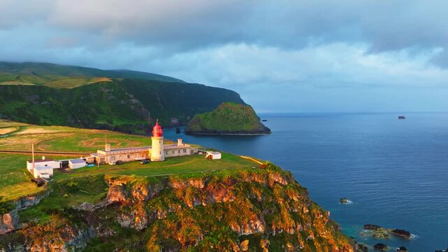 Sunrise over Farol do Albarnaz lighthouse on Flores Island, Azores, Portugal. Volcanic coast, patchwork green fields, cliffs and Atlantic ocean. Golden hour. Aerial view. Orbiting