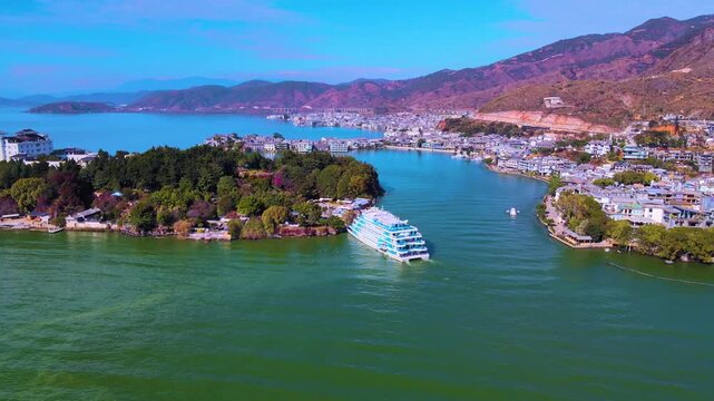 Aerial view of a multi-deck tourist cruise ship navigating Erhai Lake near Xiaoputuo Island, Dali, with traditional white-walled buildings, lush greenery, and mountainous backdrop under clear blue sky