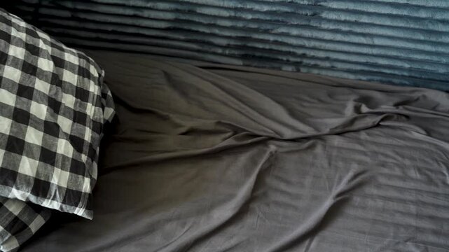 Black and white checkered pillow lying on a rumpled grey bed sheet. Soft natural light falls across the crinkled fabric of the unmade bed, concept of home comfort, lazy rest and cozy bedroom interior