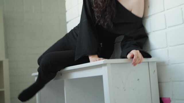 Woman dressed in black is seated indoors against brick wall. Woman adjusts positions while resting one arm on edge of white shelf with her legs swinging and feet occasionally dangling