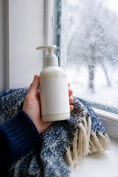 Hand holding a white lotion pump bottle with blank label on a windowsill with frost. Winter skincare product mockup on a wool blanket beside an icy window