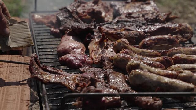 Grill master turning several pieces of meat on rural asado with fire in background, slow motion