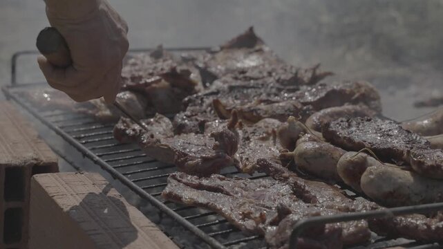 Hand flipping piece of meat on countryside asado grill with chorizo and fire, slow motion