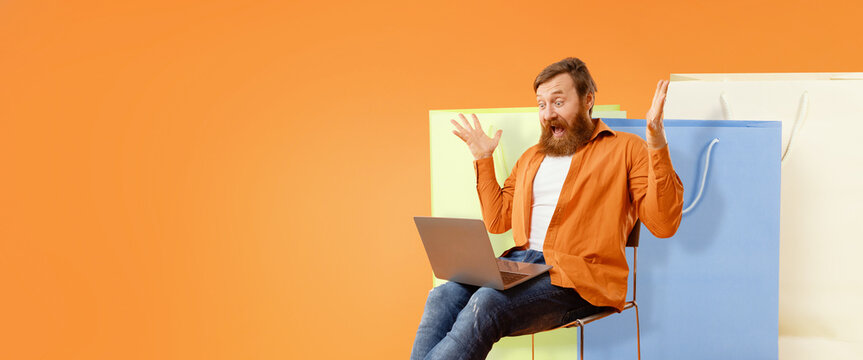 Excited Caucasian Man With Beard Sitting On Chair While Shopping Online With Laptop, Surrounded By Colorful Shopping Bags In Bright Background. Enjoying Modern Online Shopping Experience, Business