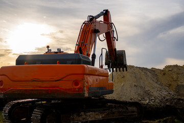 An orange tracked excavator raises its hydraulic boom and toothed bucket beside a mound of earth at a construction site under a cloudy sky. © Marina