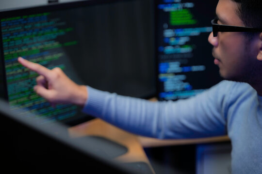 Computer programmer works at a multi monitor workstation in a dark office, reviewing programming code for software development, technical analysis, and system monitoring.